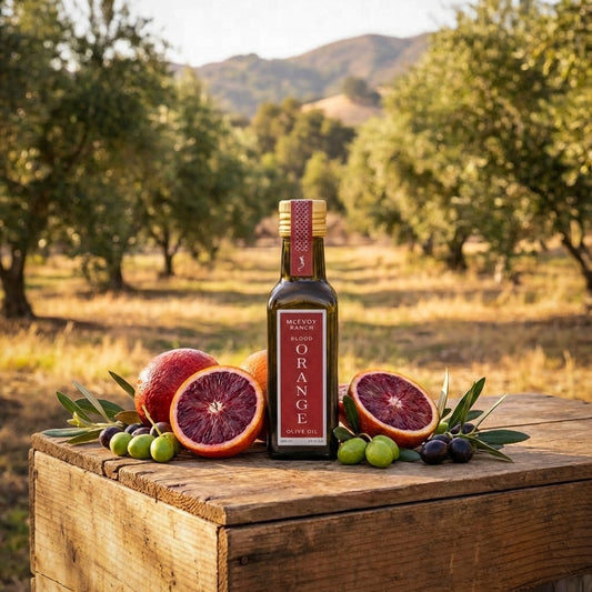 Bottle of Meevou Ranch Blood Orange Olive Oil on a wooden surface with blood oranges and olives in an outdoor setting.