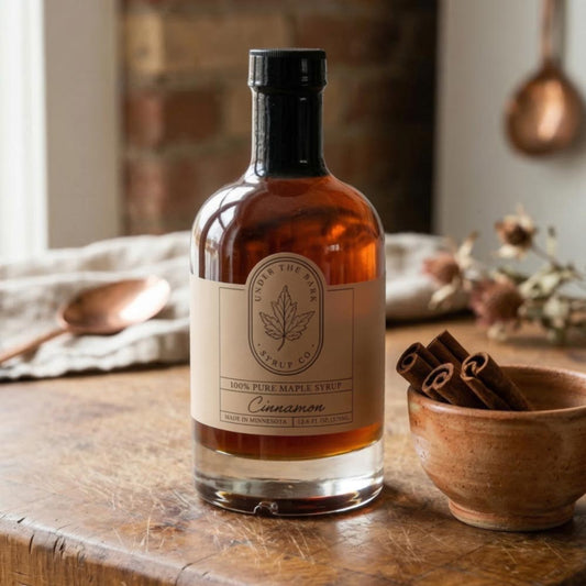 Bottle of Under The Bark cinnamon maple syrup on a wooden table with a bowl of cinnamon sticks.