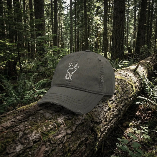Dark gray cap with a white bugling elk embroidery on a log in a forest setting