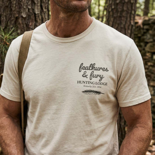 Person wearing a beige t-shirt with 'feathers & fury Hunting Lodge' design in a forest setting