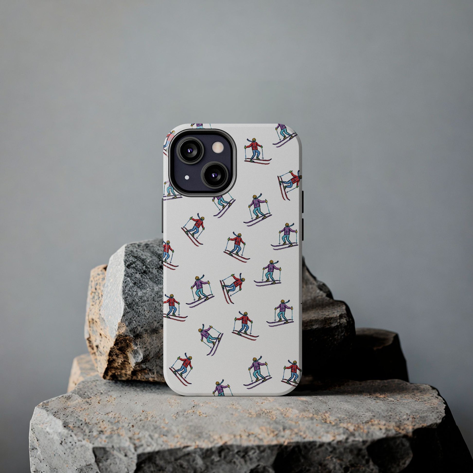 Lifestyle shot of a cell phone case with illustrated skiers resting on rugged rocks, highlighting its outdoor-inspired skiing pattern and durable design.