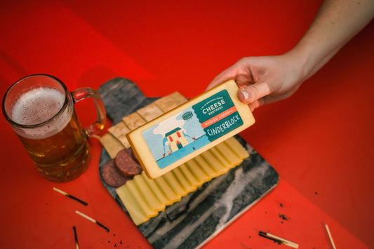 Person holding a block of smoked cheddar cheese with the Cheese Brothers cinderblock label, next to a glass of beer and cheese board on a red surface.