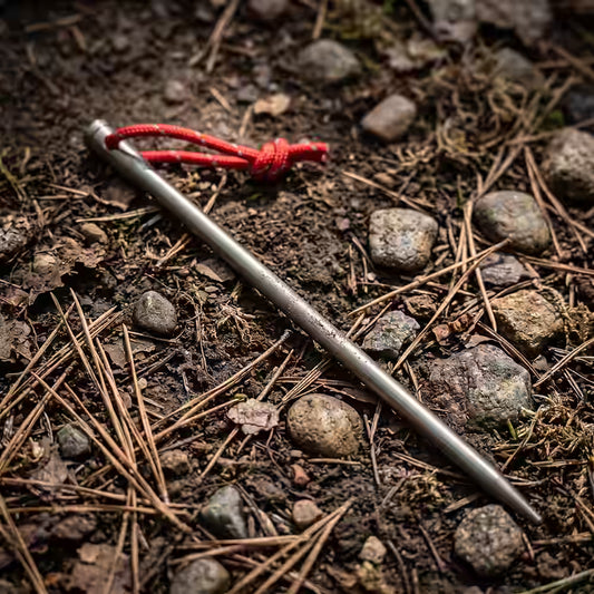 Titanium Metal tent spike with a red string tied around it lying on a forest floor covered in pine needles, soil, and small rocks.