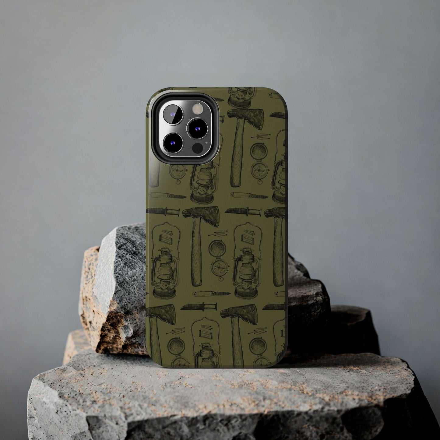 Vintage Survivalist Phone Case displayed on a rock, showcasing its rugged outdoor-inspired design with lantern, axe, knife, matches, and compass illustrations.