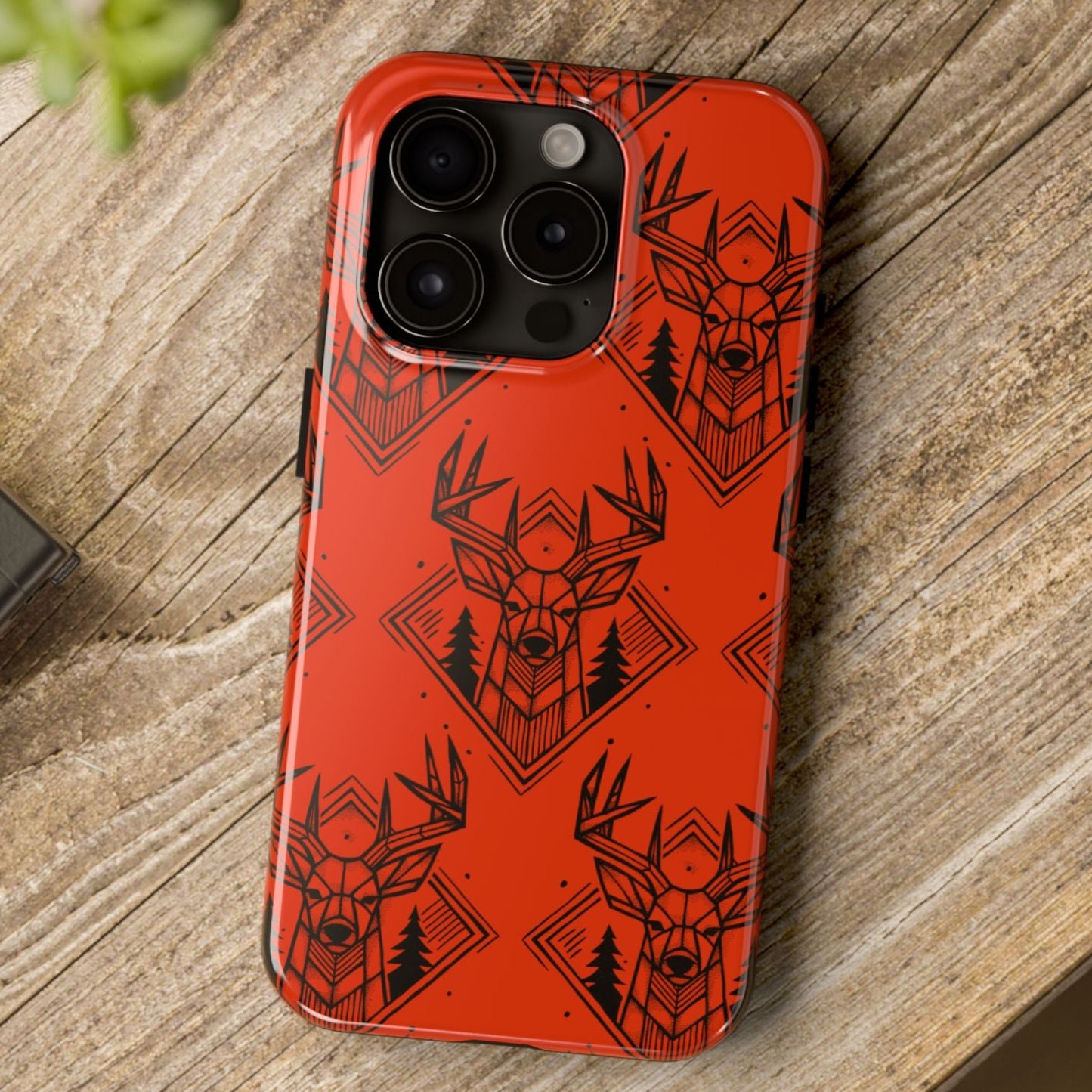 Top view on a desk of Antler Buck pattern iPhone Case in orange, featuring a bold antler and buck design for hunters. Buck is framed by pine trees and geometric shape with hand drawn design.