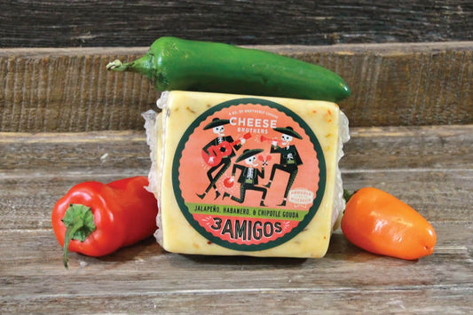 Jalapeno, habanero, chipotle gouda cheese block with 'Three Amigos' label surrounded by peppers on a wooden surface by the Cheese Brothers.