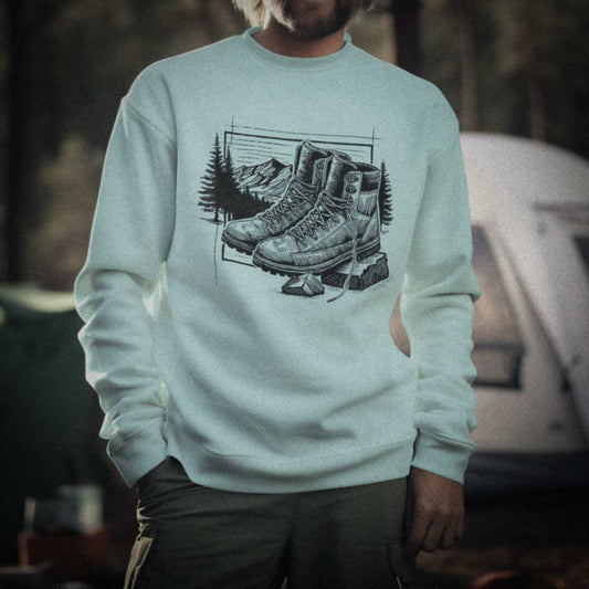 Outdoor lifestyle photography of a person at a campsite wearing the Hiking Boots Artist Series sweatshirt. Features original fine-line studio art on 100% natural cotton with no synthetic blends.