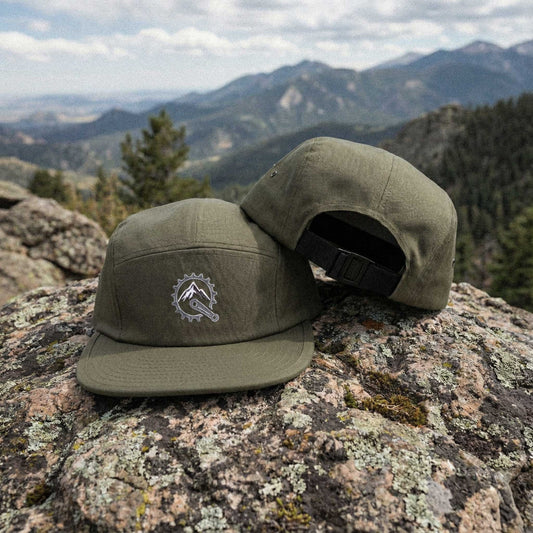 Two olive green panel caps with embroidered Mountain Bike Sprocket / Mountain Peak graphic on a rocky surface with mountains in the background by Kempain Family Outfitters.