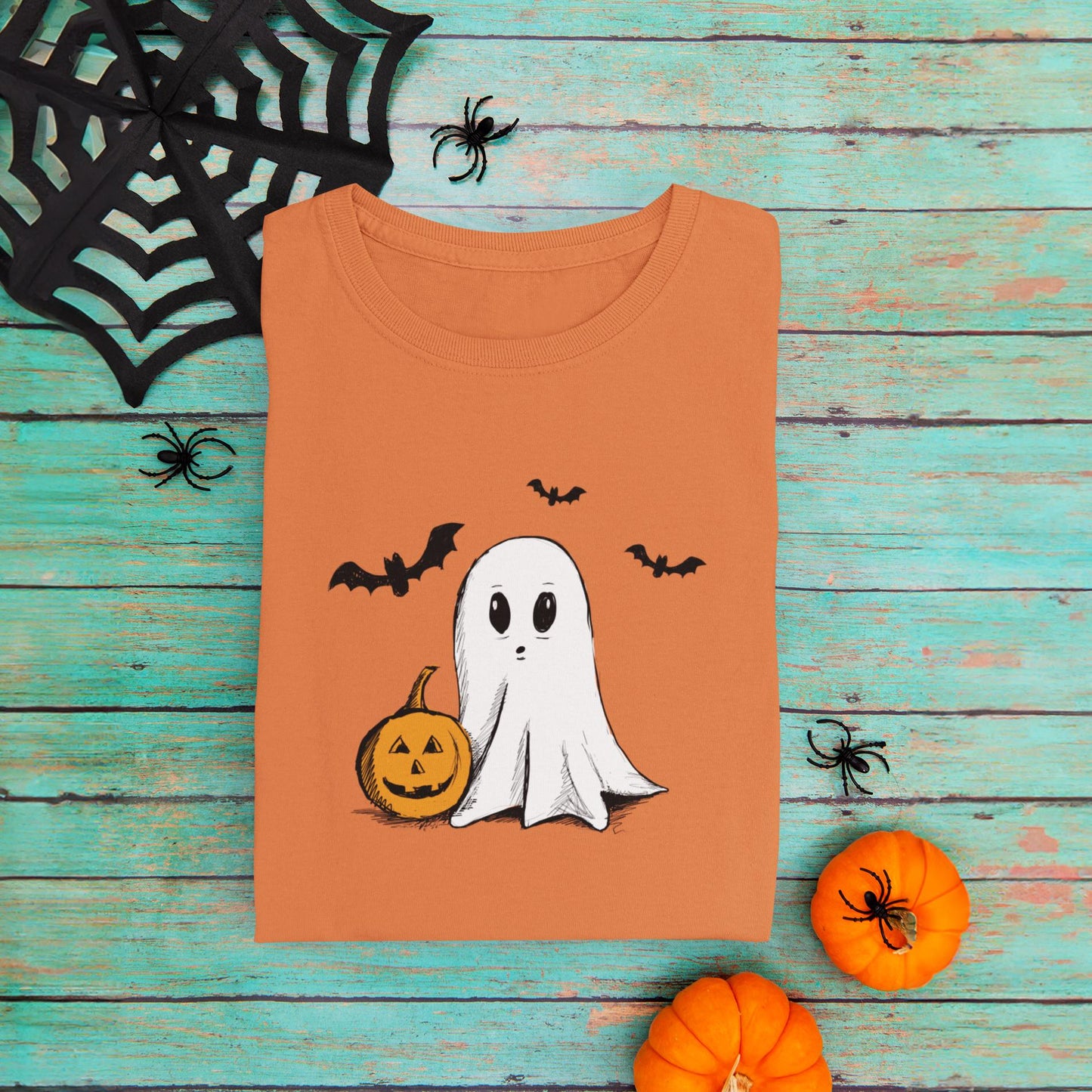 Orange Halloween shirt folded on a rustic wooden background, surrounded by Halloween items like plastic spiders, a spider web, and a pumpkin, creating a festive spooky atmosphere.