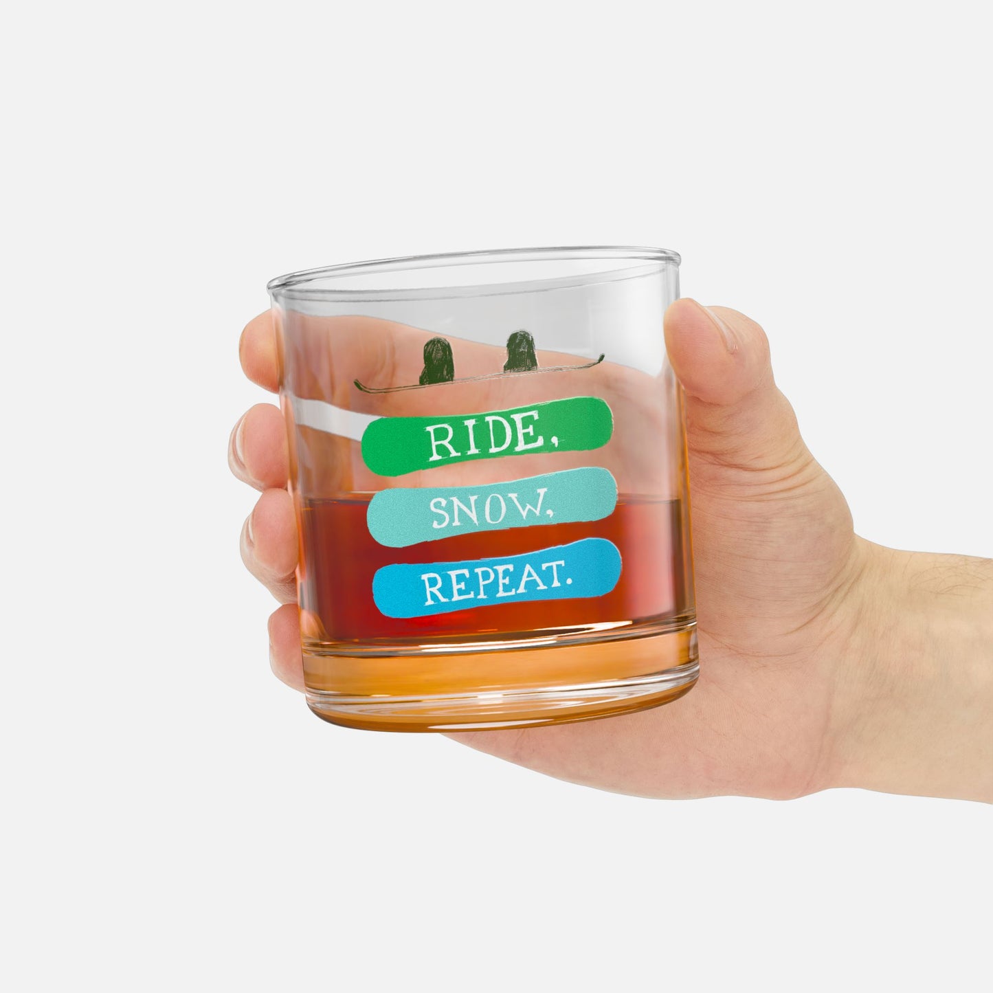 Hand holding a rocks glass with the phrase 'Ride, Snow, Repeat' and a snowboarding-themed design, perfect for relaxing after a day on the slopes.