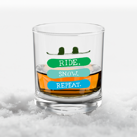 Close-up of a rocks glass with the phrase 'Ride, Snow, Repeat' and a snowboarding-themed design, displayed in a snowy pile. Perfect for snowboard enthusiasts and winter-inspired drinkware.