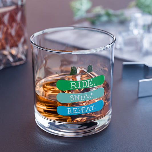Rocks glass filled with a whiskey-colored beverage, featuring the phrase 'Ride, Snow, Repeat' and a snowboarding-themed design, placed on a rustic table.