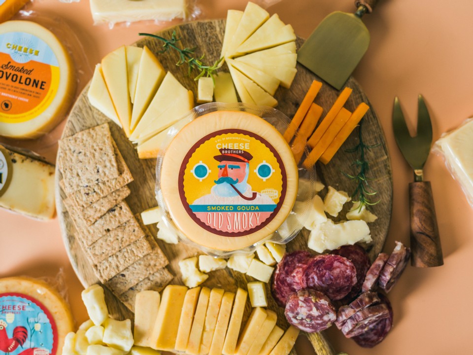 Assorted cheese platter with various types of cheese, crackers, and meats on a wooden board featuring "Old Smoky" Smoked Gouda by the Cheese Brothers in the middle.