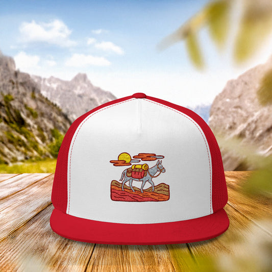 Trail Legends Pack Mule Trucker Cap resting on a wooden table with a rugged mountain backdrop, highlighting the hat’s outdoor-ready design and adventurous spirit with embroidered design.