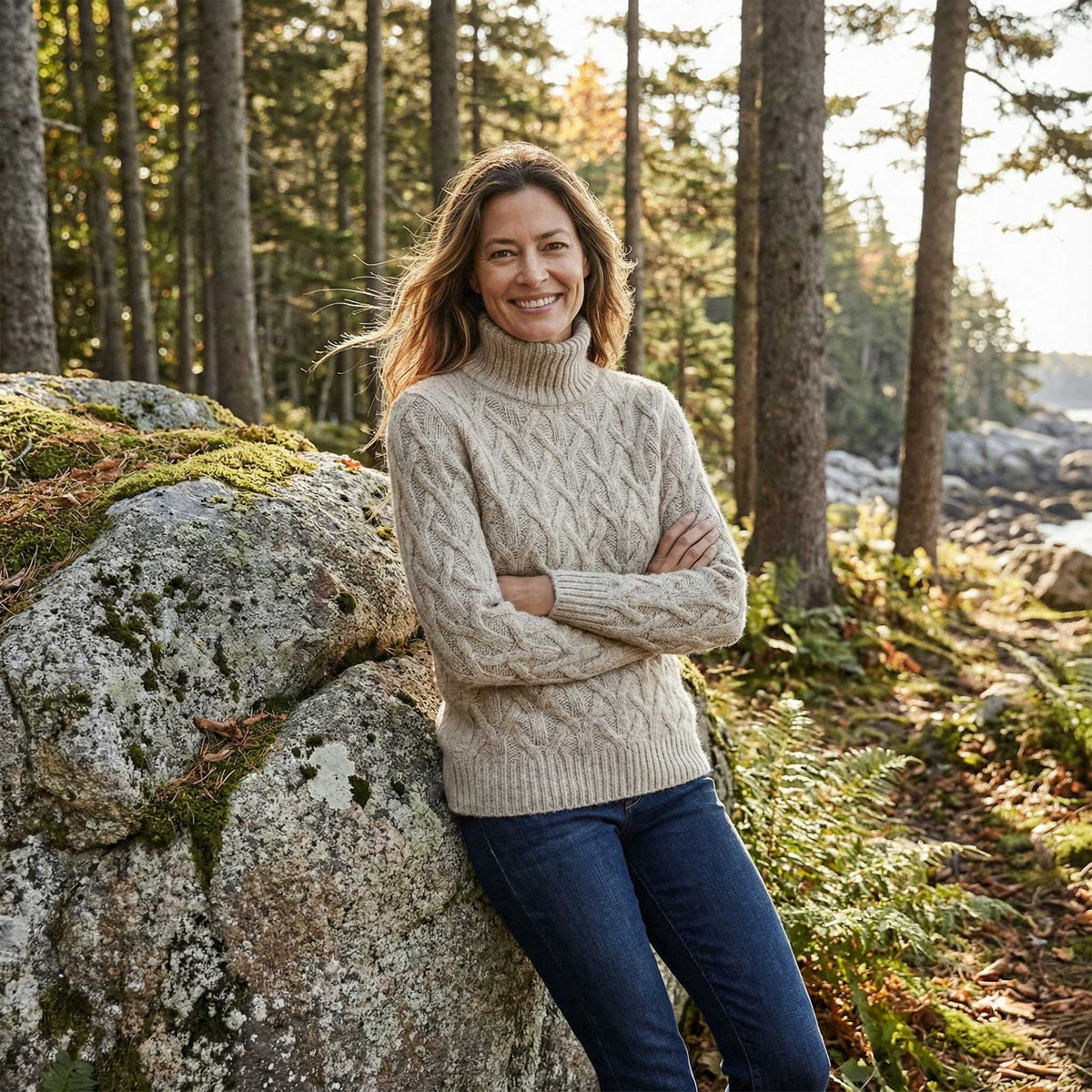 Woman in a beige Aplaca sweater and blue jeans standing in a forest with trees and rocks in the background.