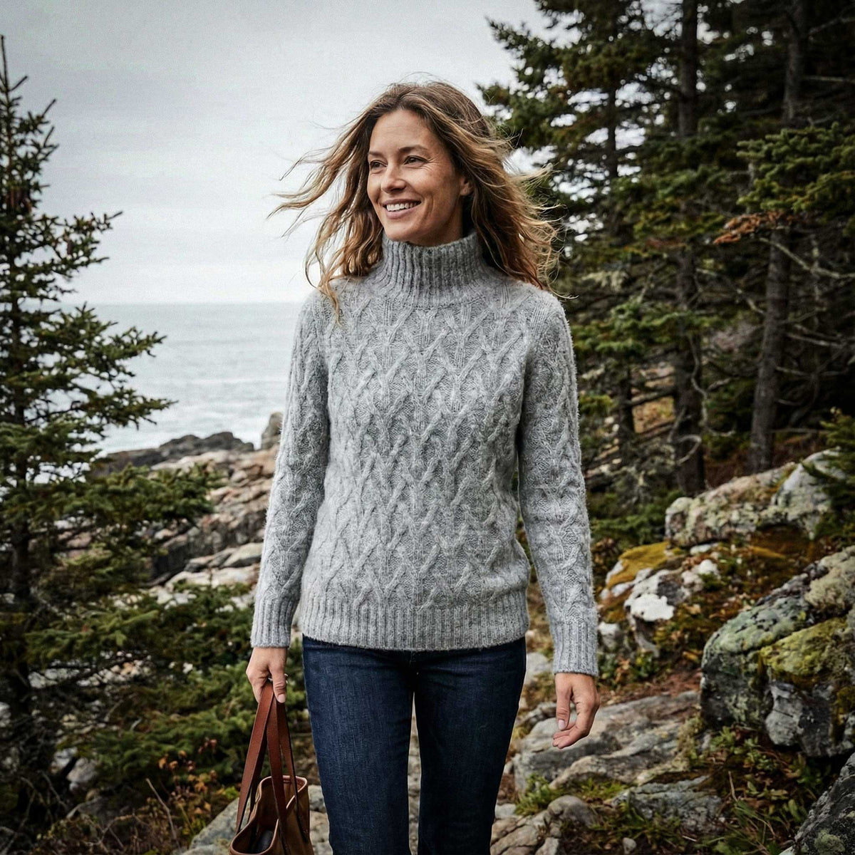 Woman wearing a gray sweater standing in a forested area with trees and rocks in the background.