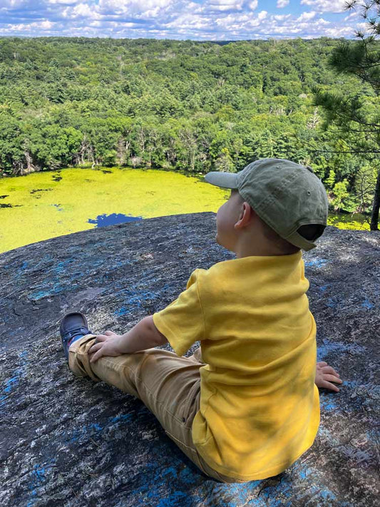 A young boy wearing a yellow t-shirt, khaki pants, and an olive green baseball cap sits on a rocky overlook 'Ross's Cliff", looking out over a lush green forest and a bright green pond under a cloudy sky at Old Furnace State Park.