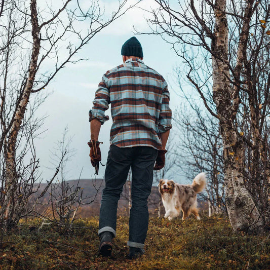 A man wearing a thick flannel shirt with his dog in the wilderness carrying and axe.