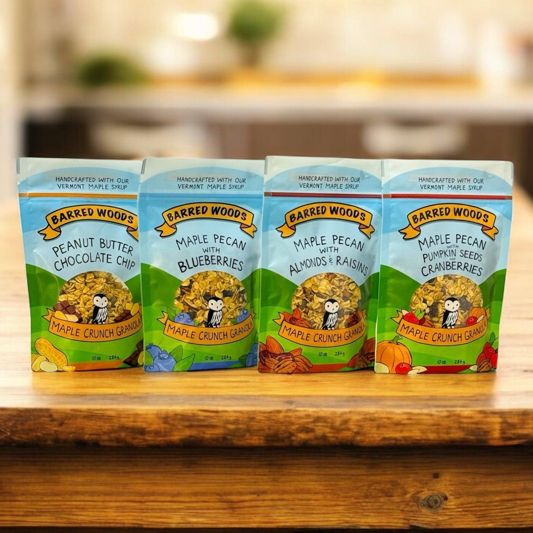 Four packages of Barred Woods Maple Crunch Granola on a wooden surface with a blurred kitchen background.