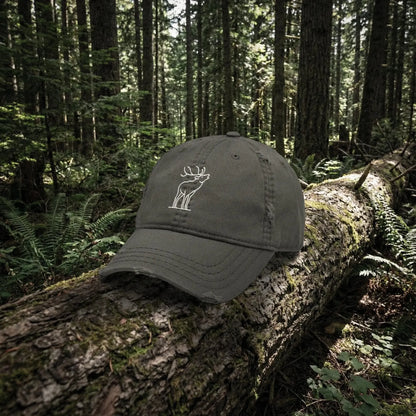 Dark gray cap with a white bugling elk embroidery on a log in a forest setting