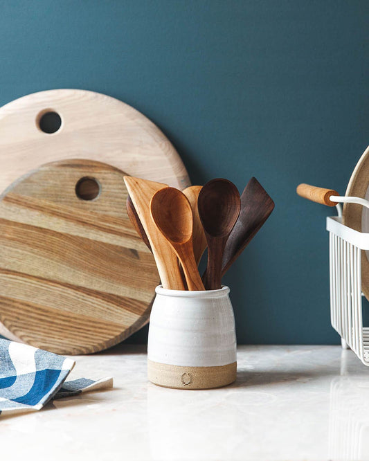 Wooden spoons in a white wheel thrown crock container with a cutting board and kitchen towel on a blue wall background