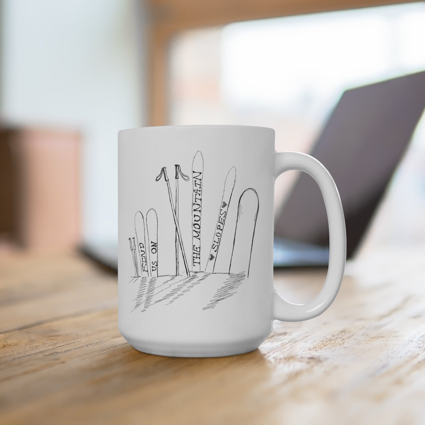 Front view on a desk of this 15oz ceramic mug is perfect for anyone who loves the thrill of the slopes and cozy mornings. Featuring a minimalist pen-and-ink illustration of skis and a snowboard stuck in snow, accompanied by the hand-written phrase Find Us on the Mountain Slopes, this mug captures the essence of winter adventures. 