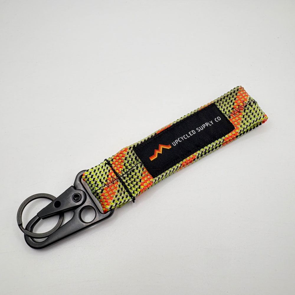 A green and orange braided climbing rope recycled fabric keychain with a black metal carabiner and a black label reading 'UPCYLED SUPPLY CO' on a grey background.