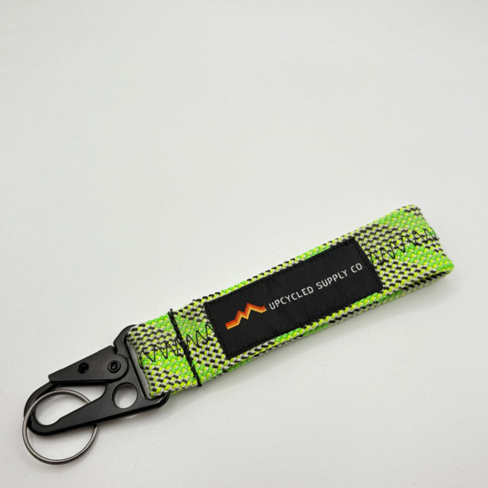 A green braided climbing rope recycled fabric keychain with a black metal carabiner and a black label reading 'UPCYLED SUPPLY CO' on a grey background.