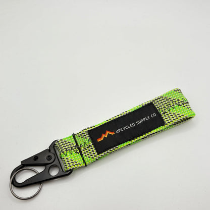 A green braided climbing rope recycled fabric keychain with a black metal carabiner and a black label reading 'UPCYLED SUPPLY CO' on a grey background.