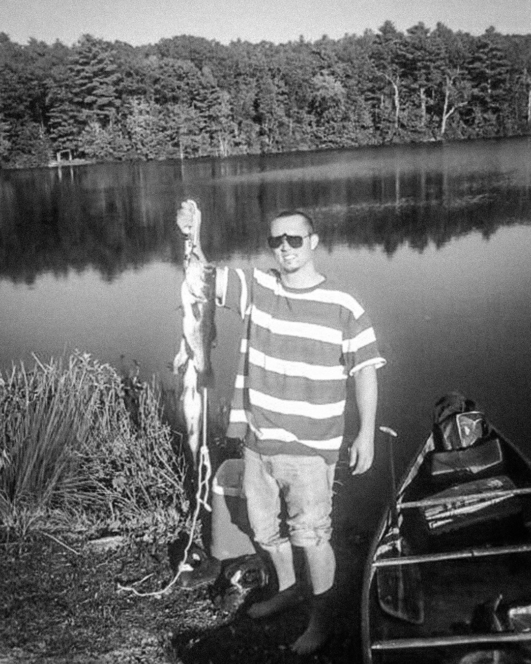 A black and white photo of Jordan Kempain standing next to a canoe at a lake holding a stringer with two Largmouth Bass that he cought.