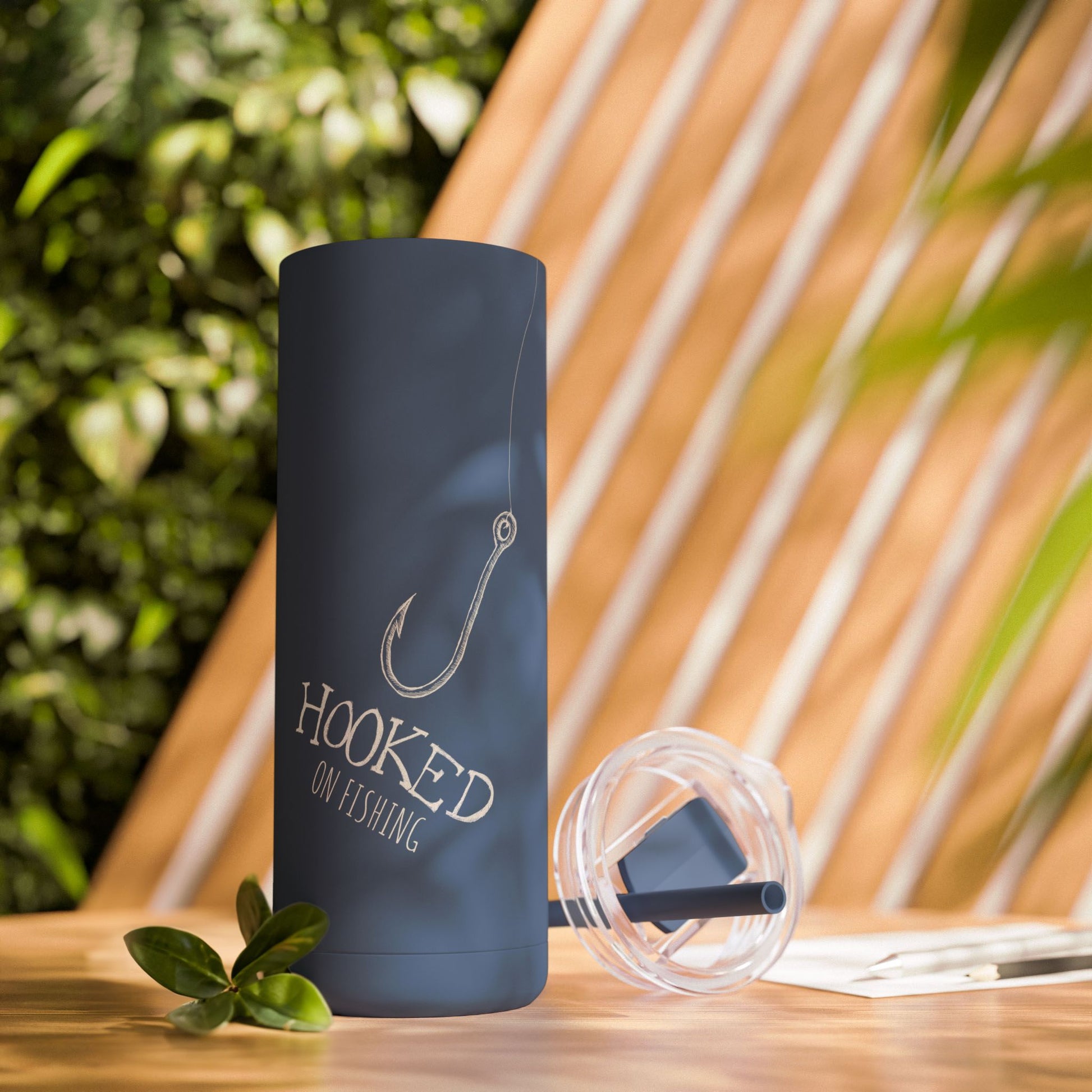 A lifestyle image of Hooked on Fishing Blue Tumbler - Personalized Gift with Fish Hook & Line Design showed on a table with cover and straw off outdoors.