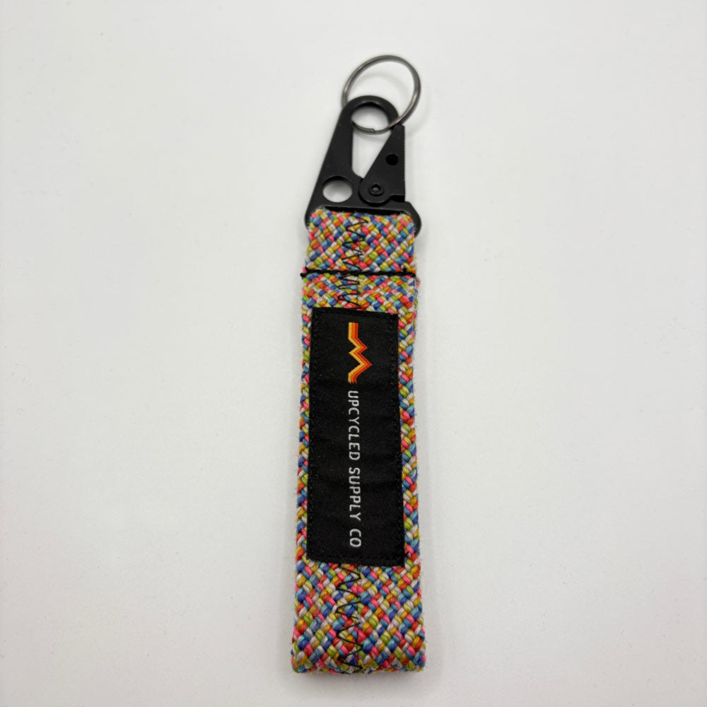 A multicolor braided climbing rope fabric keychain with a black metal carabiner and a black label reading 'UPCYLED SUPPLY CO' laying flat on a grey background.