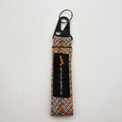 A multicolor braided climbing rope fabric keychain with a black metal carabiner and a black label reading 'UPCYLED SUPPLY CO' laying flat on a grey background.
