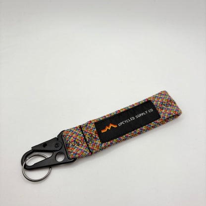 A multicolor braided climbing rope fabric keychain with a black metal carabiner and a black label reading 'UPCYLED SUPPLY CO' on a gray background.