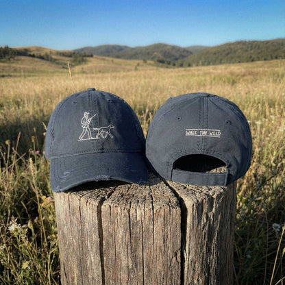Lifestyle photograph of Front and back view of navy blue "The Pointer Cap" featuring embroidered Hunter with Dog on front and "Walk the Wild" text on back on 100 percent cotton twill distressed dad cap by Kempain Family Outfitters.