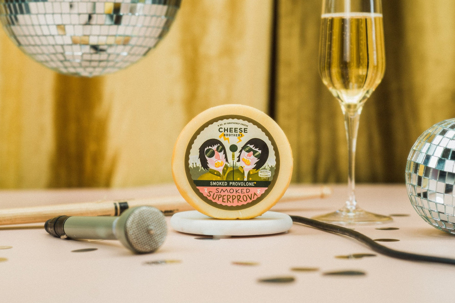 A round cheese wheel labeled 'CHEESE CLUB SMOOKED SUPERPROV' sits on a small white dish on a table, styled with a champagne flute, two disco balls, a microphone, and confetti against a warm golden backdrop.