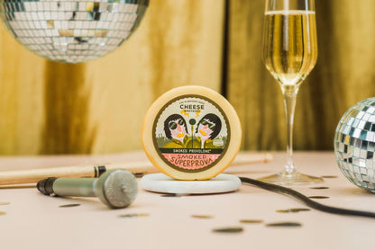A round cheese wheel labeled 'CHEESE CLUB SMOKED SUPERPROVA' sits on a small white dish on a table, styled with a champagne flute, two disco balls, a microphone, and confetti against a warm golden backdrop.