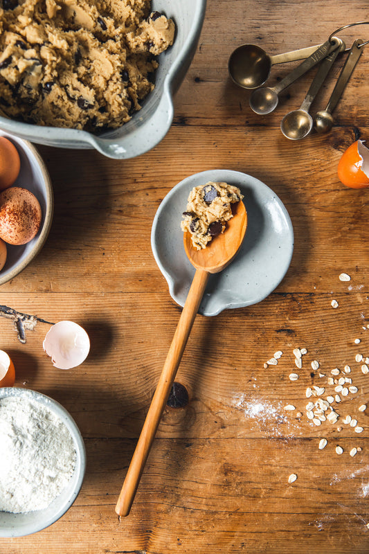 Bowl of cookie dough with a wooden spoon on a wooden table with ingredients and utensils.