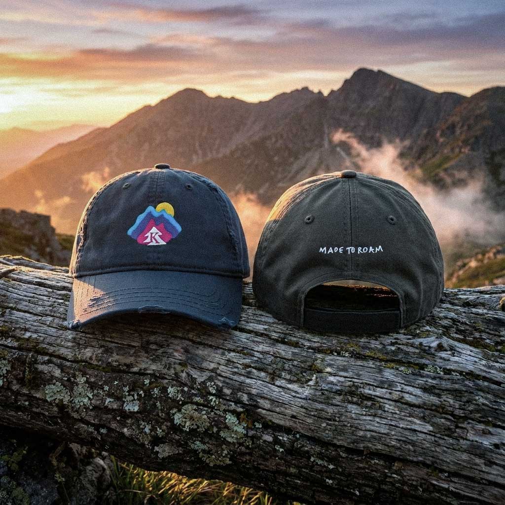 Two 100 percent cotton twill caps with Made To Roam embroidered graphics on a log with a mountainous landscape in the background during sunset by Kempain Family Outfitters.