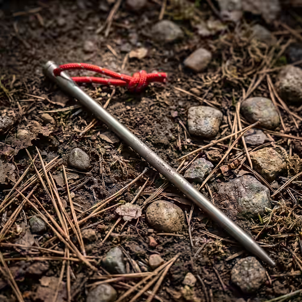 Titanium Metal tent spike with a red string tied around it lying on a forest floor covered in pine needles, soil, and small rocks.