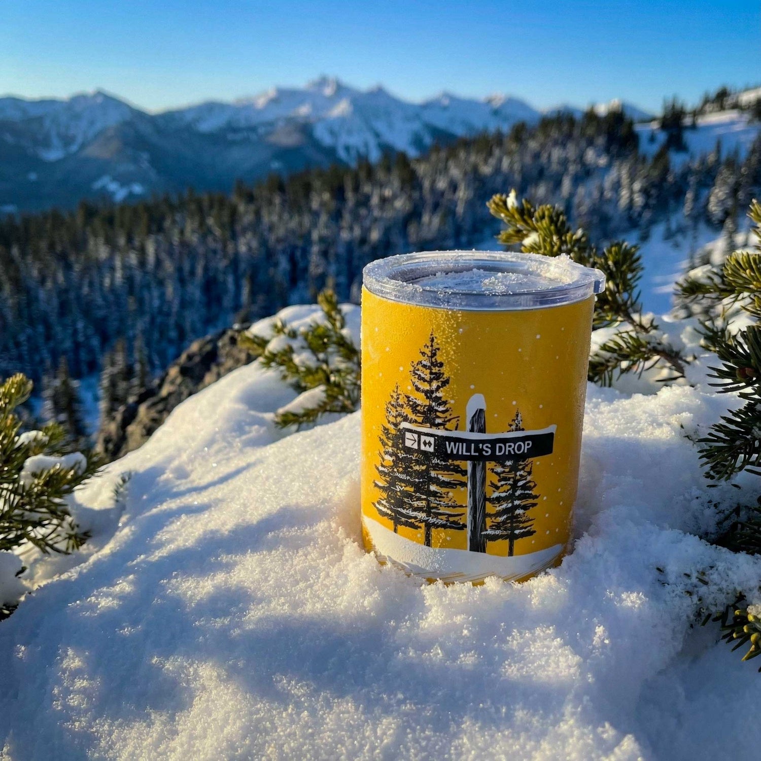 Custom tumbler featuring a ski trail sign with yellow base tumbler and custom tex "Will's Drop" with double black diamond symbol and a tree design in the snow, with mountains in the background