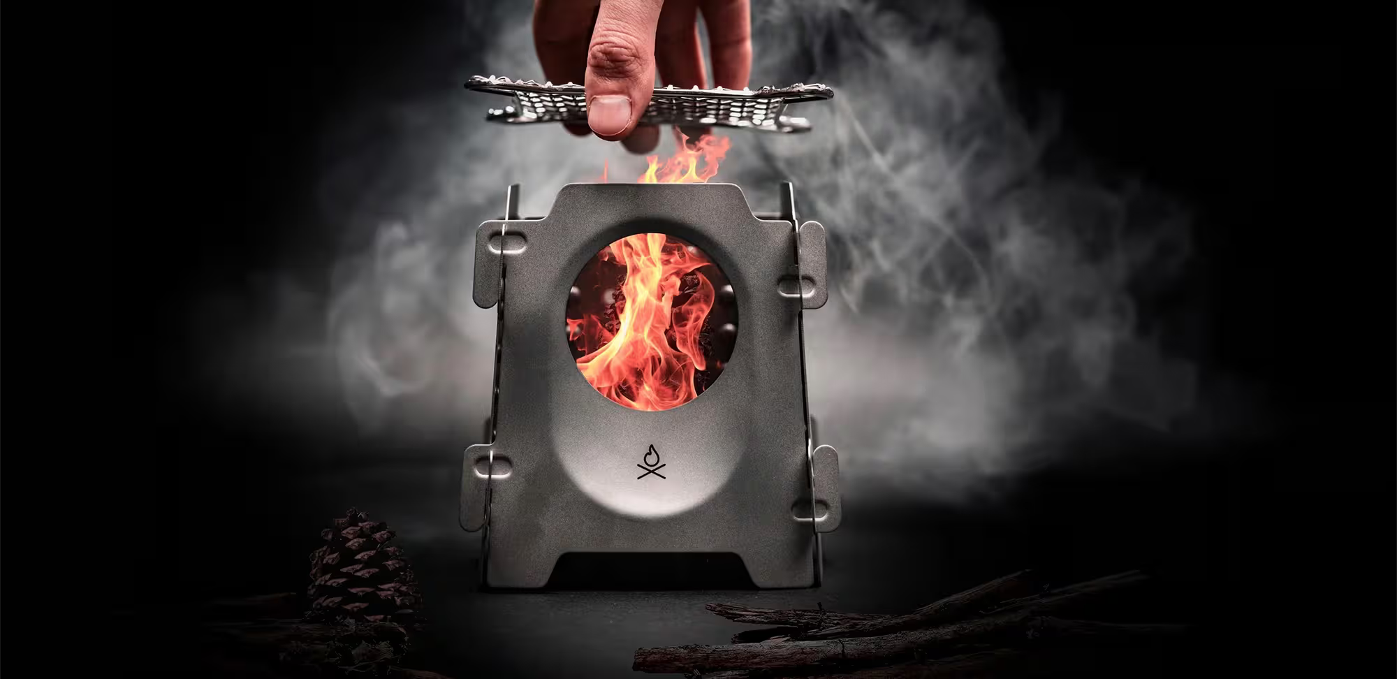 Hand holding a cooking grate above a small lit camping stove with flames, on a dark background.