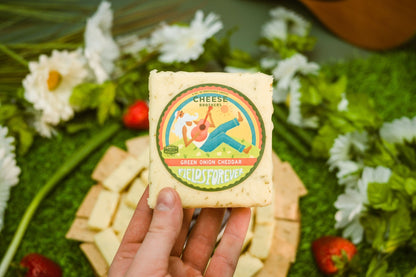 A block of Green Onion Cheddar held above a cheese board by Cheese Brothers, Inc. sold by Kempain Family Outfitters.