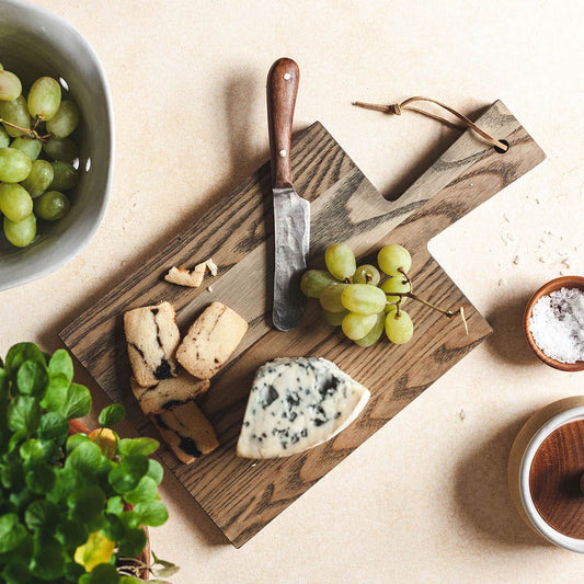 Farmhouse Pottery wooden cutting board with cheese, grapes, and a knife on a light surface.