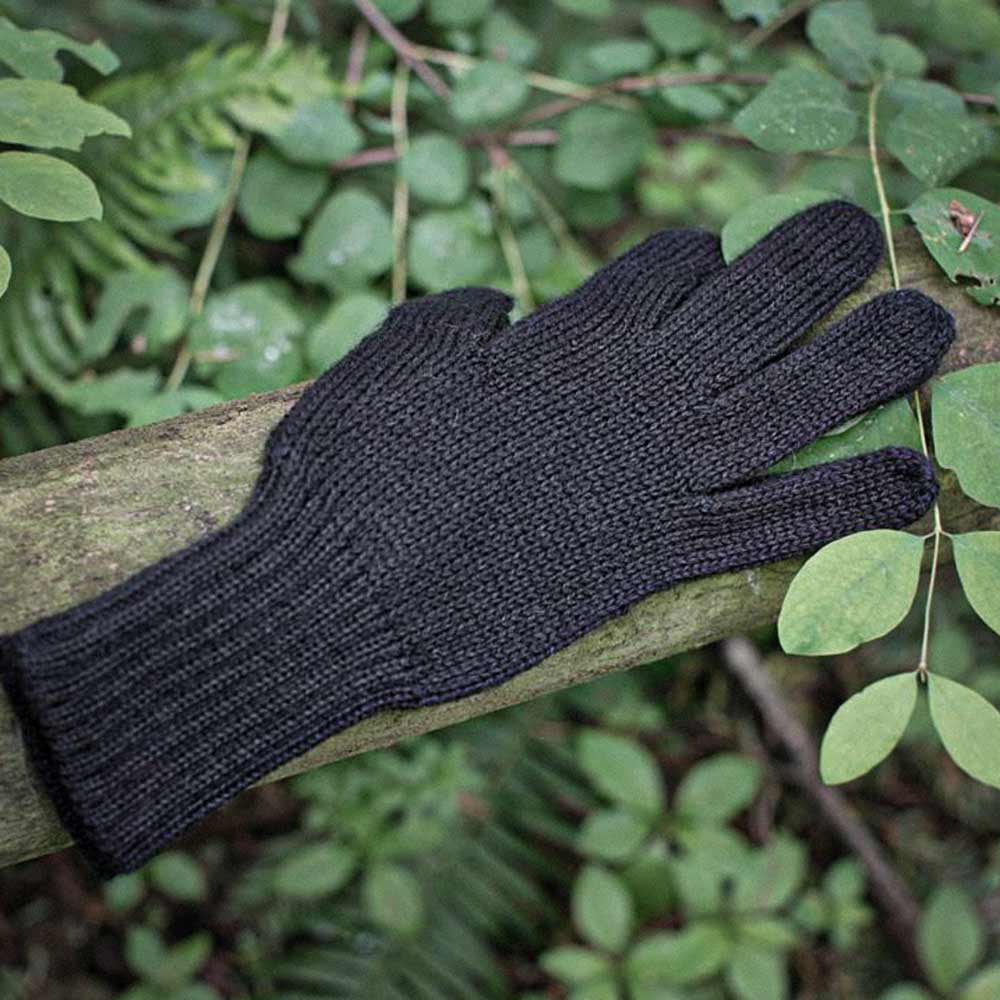 A single dark grey ribbed knit alpaca glove resting on a weathered wooden branch outdoors, surrounded by green leaves and ferns.