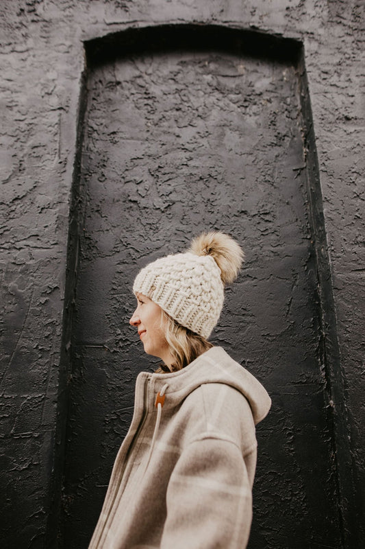 Person wearing a beige coat and knit beanie with a pom-pom against a textured black wall.