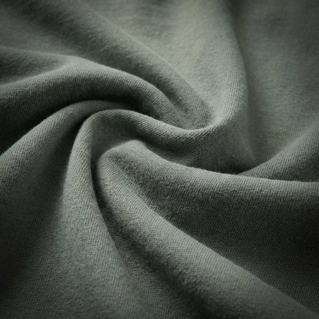 Macro detail of 100% ring-spun US cotton fabric in muted sage green, showing the heavyweight texture and natural fiber weave with zero synthetic blends.