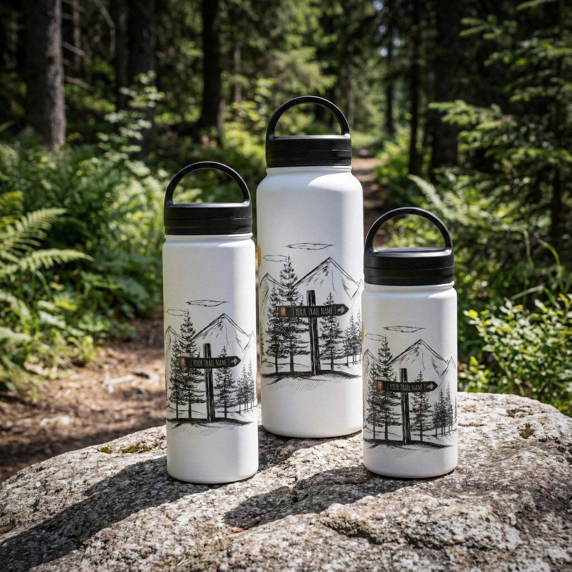 A collection of 12oz, 18oz, and 32oz custom mountain-etched water bottles arranged on a granite rock, showing the size range for family hiking trips.