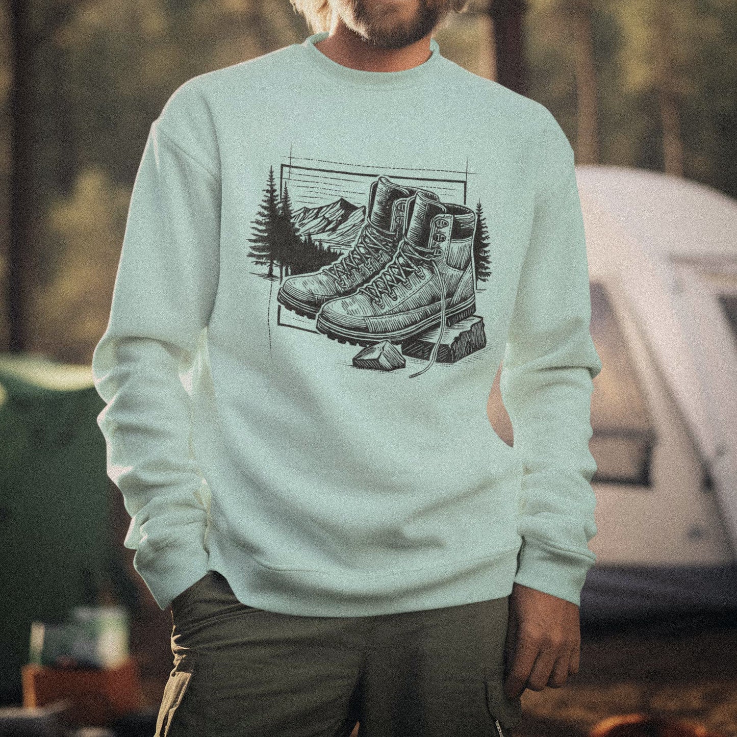 Outdoor lifestyle photography of a person at a campsite wearing the Hiking Boots Artist Series sweatshirt. Features original fine-line studio art on 100% natural cotton with no synthetic blends.