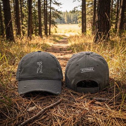 Front and back views of a dark charcoal grey distressed dad hat resting on a pine forest trail. The front features a minimalist white line-art embroidery of a jackalope, and the back features the white embroidered text "MYTHBORN".